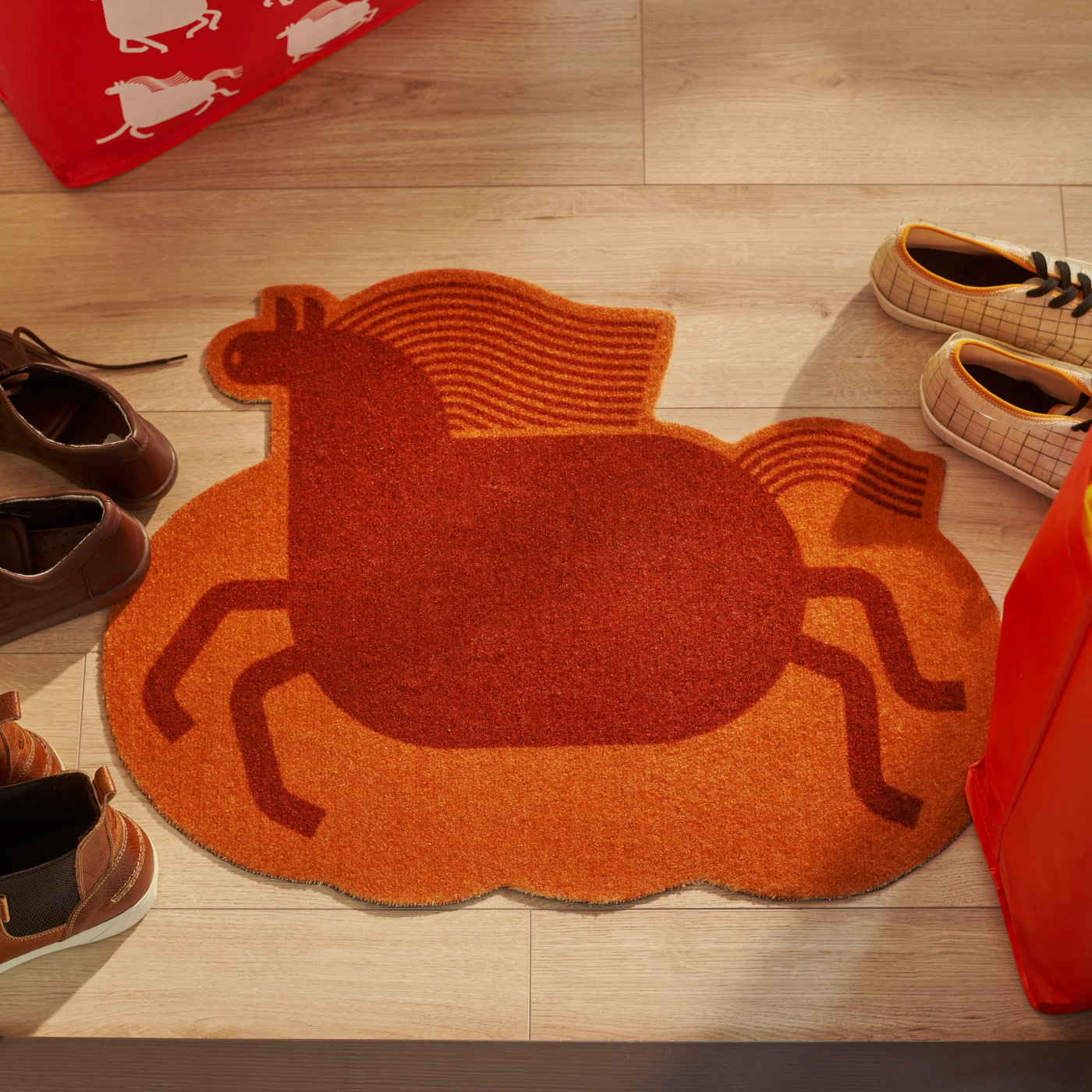 A close-up of a FÖSSTA door mat with a red horse design, placed on a light wood floor and surrounded by shoes.