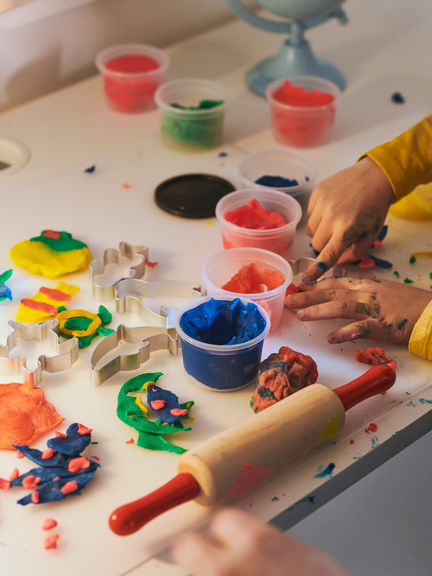 Una serie de objetos y manualidades infantiles sobre una mesa. Se ven unas manos pequeñas amasando masitas de colores.