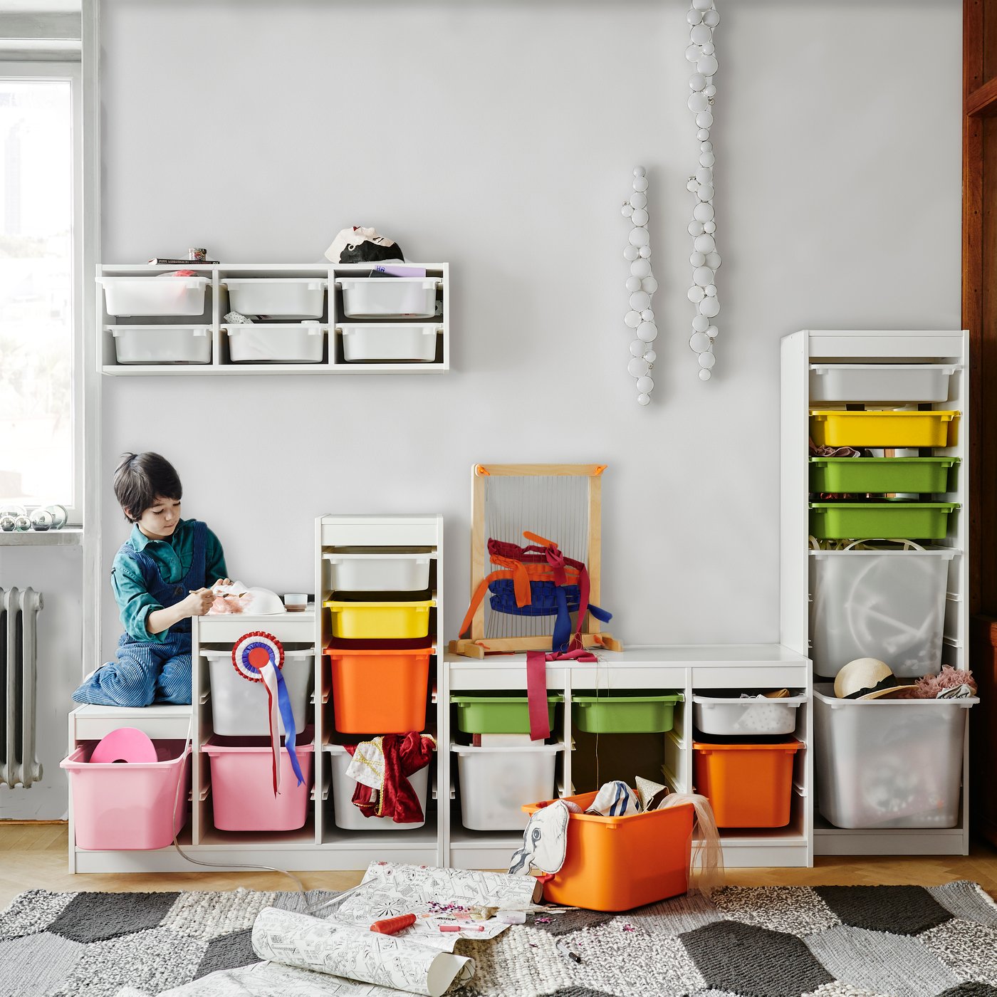 Un niño sentado sobre un mueble TROFAST de distintas alturas con cajas plásticas, grandes y pequeñas, de distintos colores. Una de las cajas está fuera del mueble y contiene artículos para hacer manualidades. 