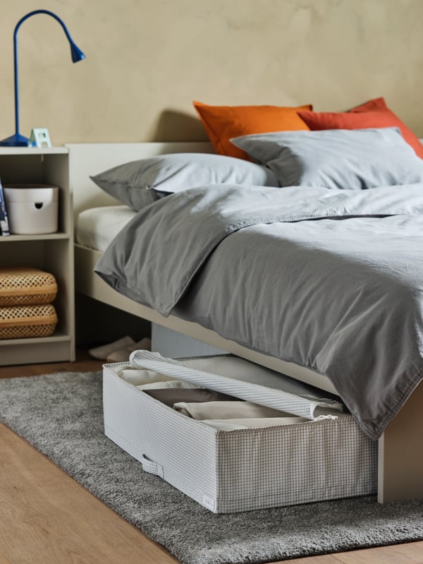 A light beige bed frame with headboard, a light beige bedside table, a white/grey storage case, a LED work lamp.