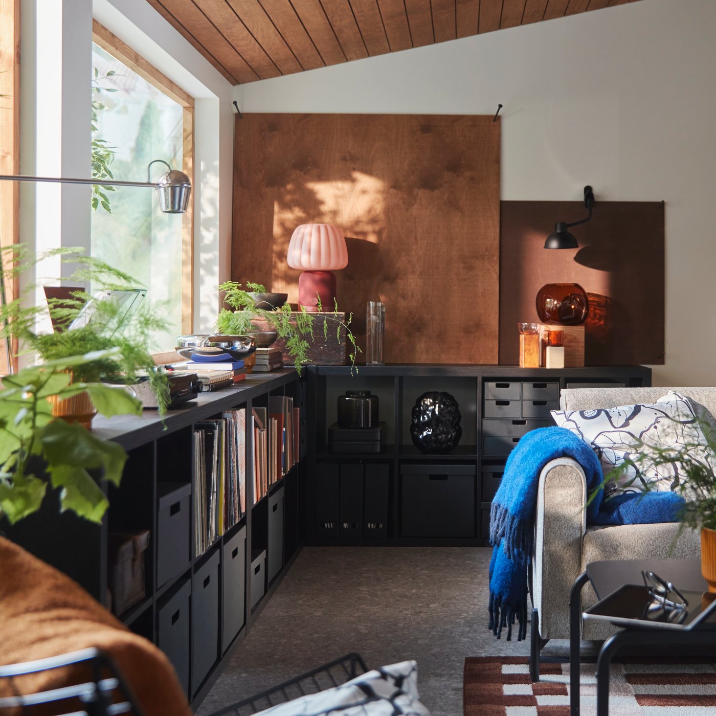 Ein Wohnzimmer mit einem schwarzbraunen KALLAX Regal, das mit Büchern und Dekorationsartikeln bestückt ist, neben einem grauen Sofa.