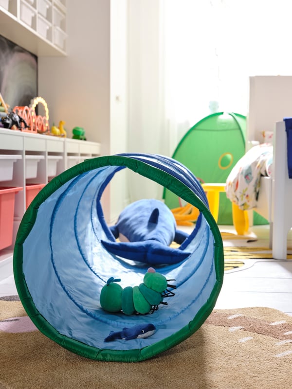 Blick in einen DVÄRGMÅS Spieltunnel, in dem ein grünes Stofftier in Raupenform sowie ein BLÅVINGAD Stoffspielzeug liegen.