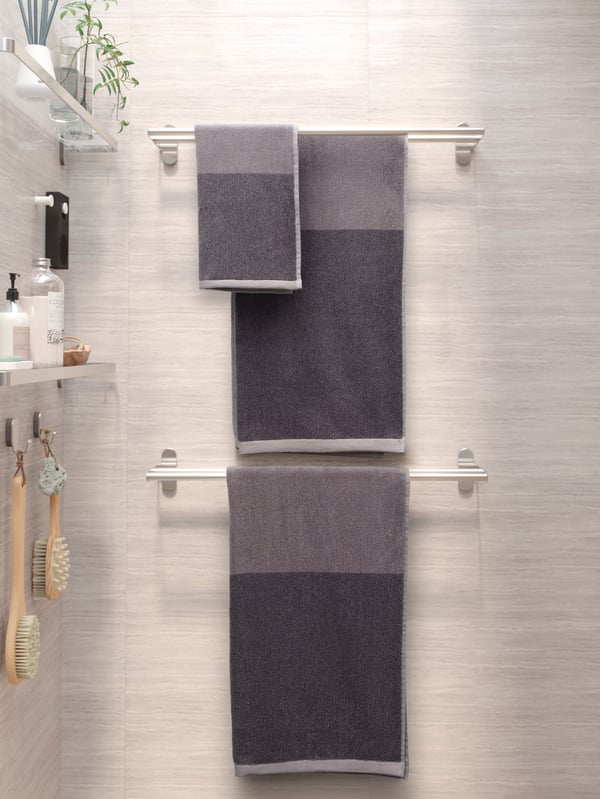 Three grey towels hanging from two BROGRUND towel rails. On the wall to the left are glass shelves with shower products.
