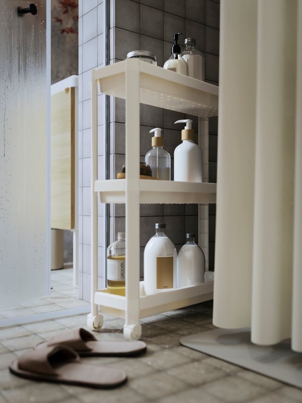 A VESKEN trolley in white holding diverse bathroom jars and containers plus a pair of SKOVELSJÖN slippers in beige.