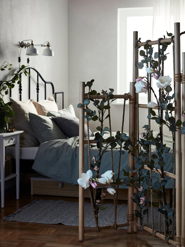 A SAGSTUA bed, HEMNES nightstand, and a plant stand with several SMYCKA artificial leaves attached to it, inside a bedroom.
