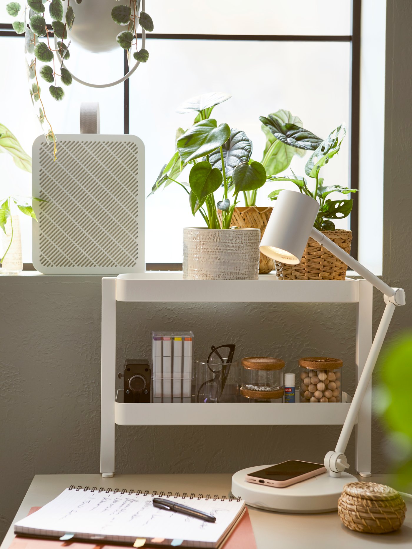 A white NYMÅNE work lamp with wireless charging on a desk, next to a window sill with plants and an UPPÅTVIND air purifier.