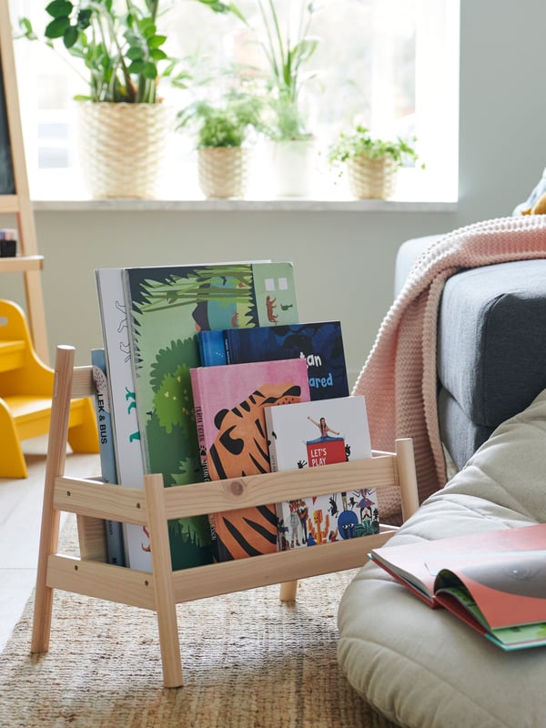 In a light-filled room, a FLISAT bookshelf containing children’s books sits on a rattan rug next to the edge of a sofa.