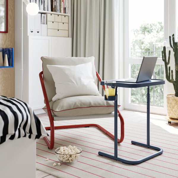 An armchair with a BJÖRKÅSEN laptop stand in blue in front of it, with a cup of juice in the holder.