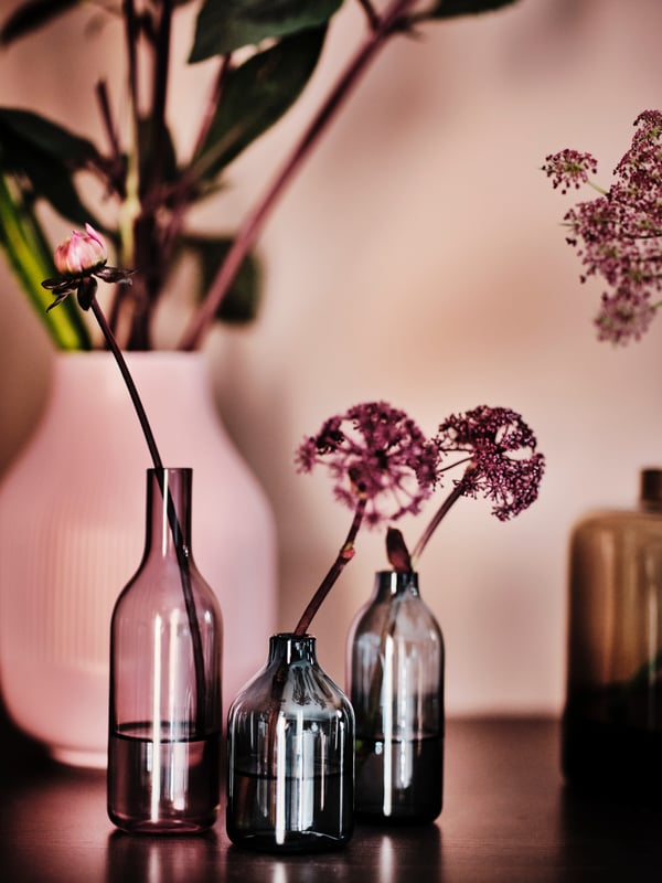Single stem flowers in lilac light grey-blue/black-blue NIMTRÄD vases, set of 3 in front of a pink GRADVIS vase of flowers.