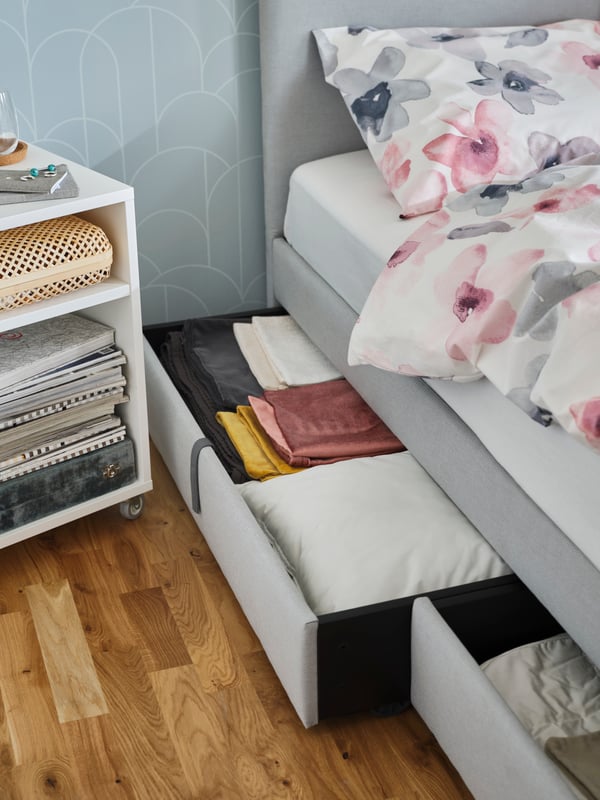 The side of a grey GLADSTAD bed showing the open under bed storage boxes filled with coloured and white textiles.