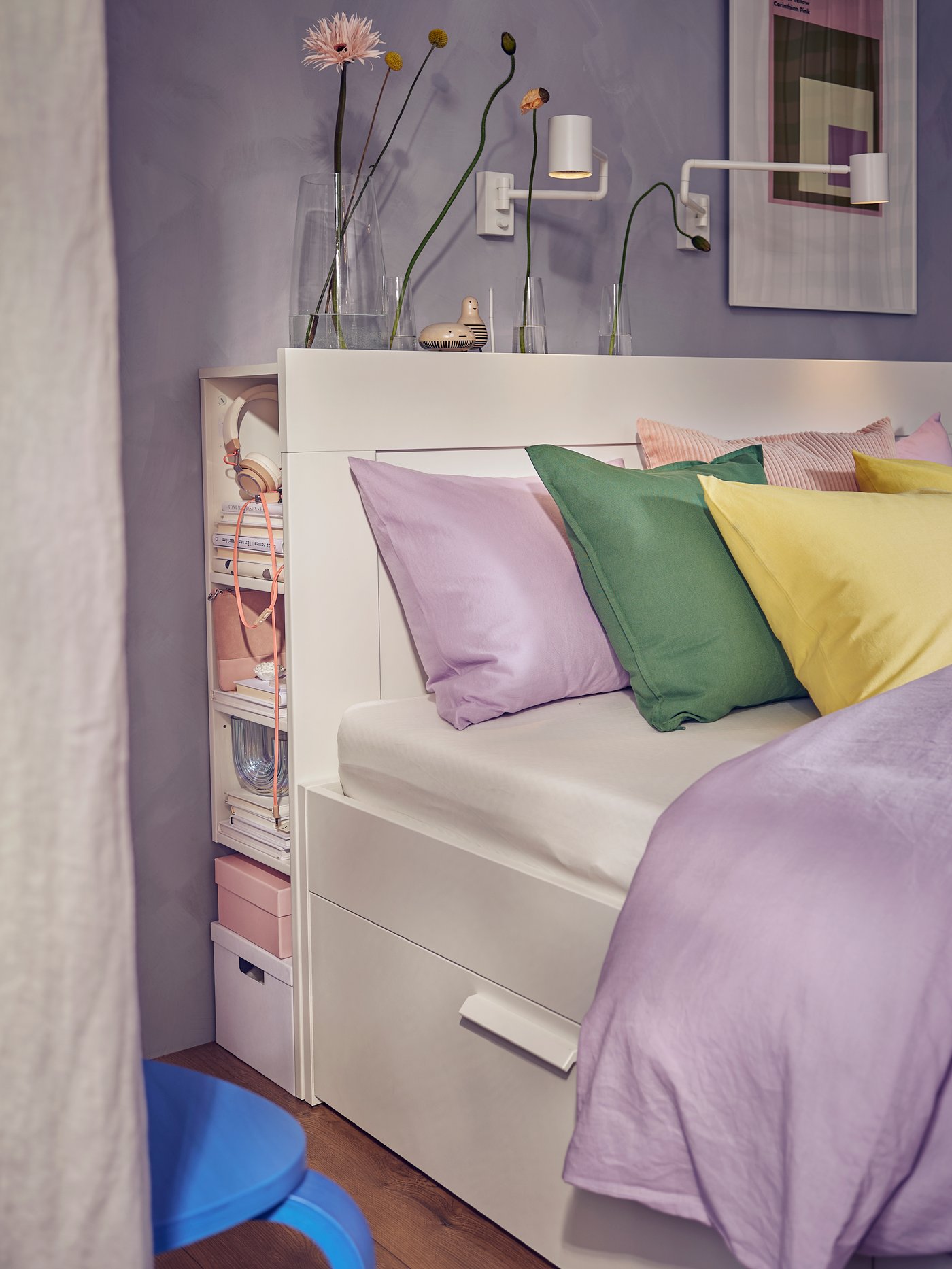 Un coin chambre soigneusement aménagé avec un lit blanc à tiroirs de rangement, des coussins pastel, une étagère à livres et des vases fleuris sur la tête de lit.