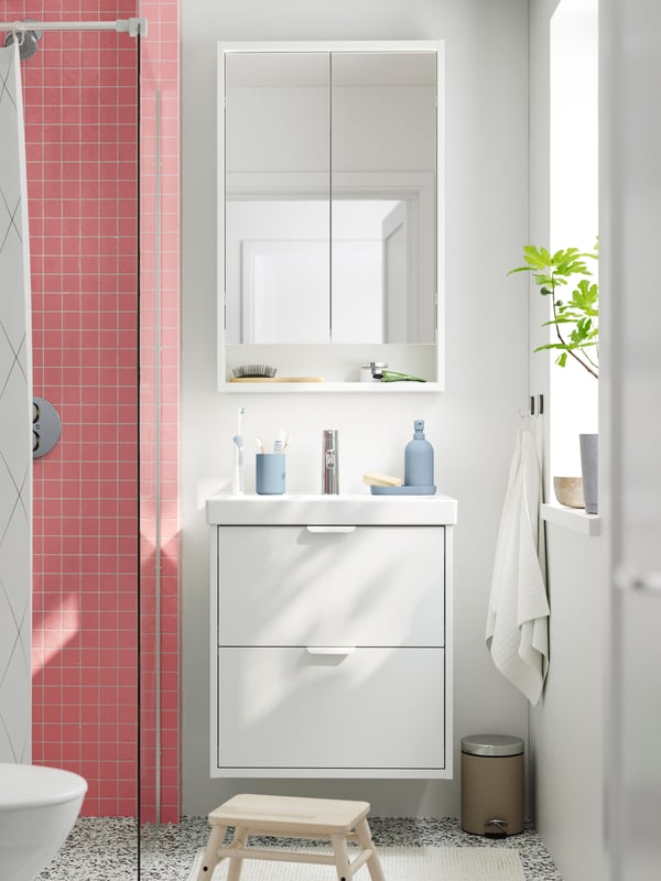 A white HAVBÄCK vanity sink with two drawers is mounted under a white mirror cabinet next to a pink-tiled shower in a bathroom.