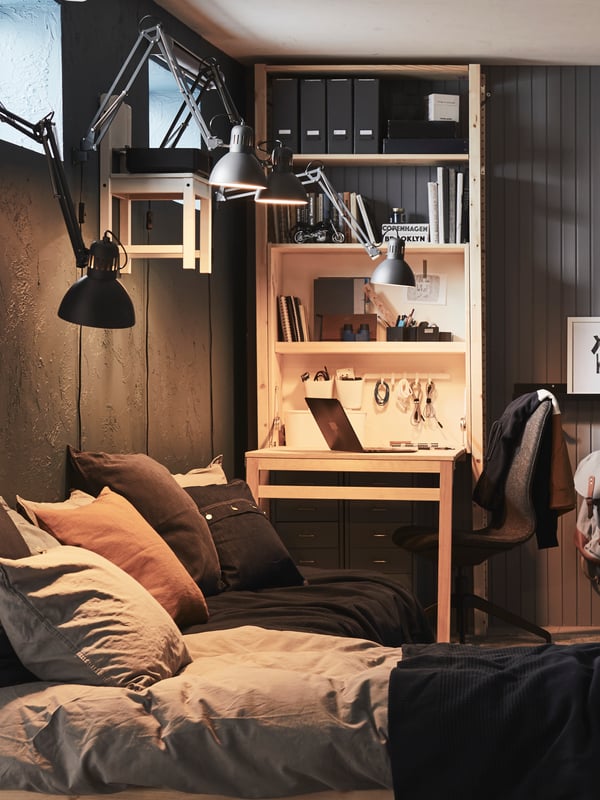 Four, dark grey, TERTIAL work lamps fixed to a wall shine down onto a bed and a pine, IVAR storage unit with foldable table.