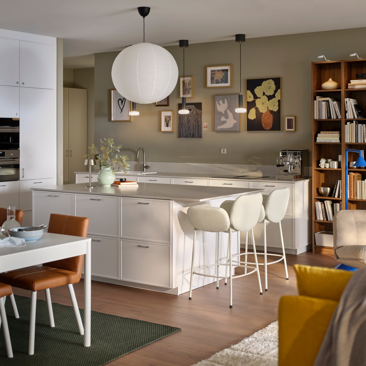 Eine moderne Küche mit weißen ASPUDDEN-Fronten, einer Kücheninsel und SYNTORP-Barhockern, die zum Wohnzimmer hin offen ist.