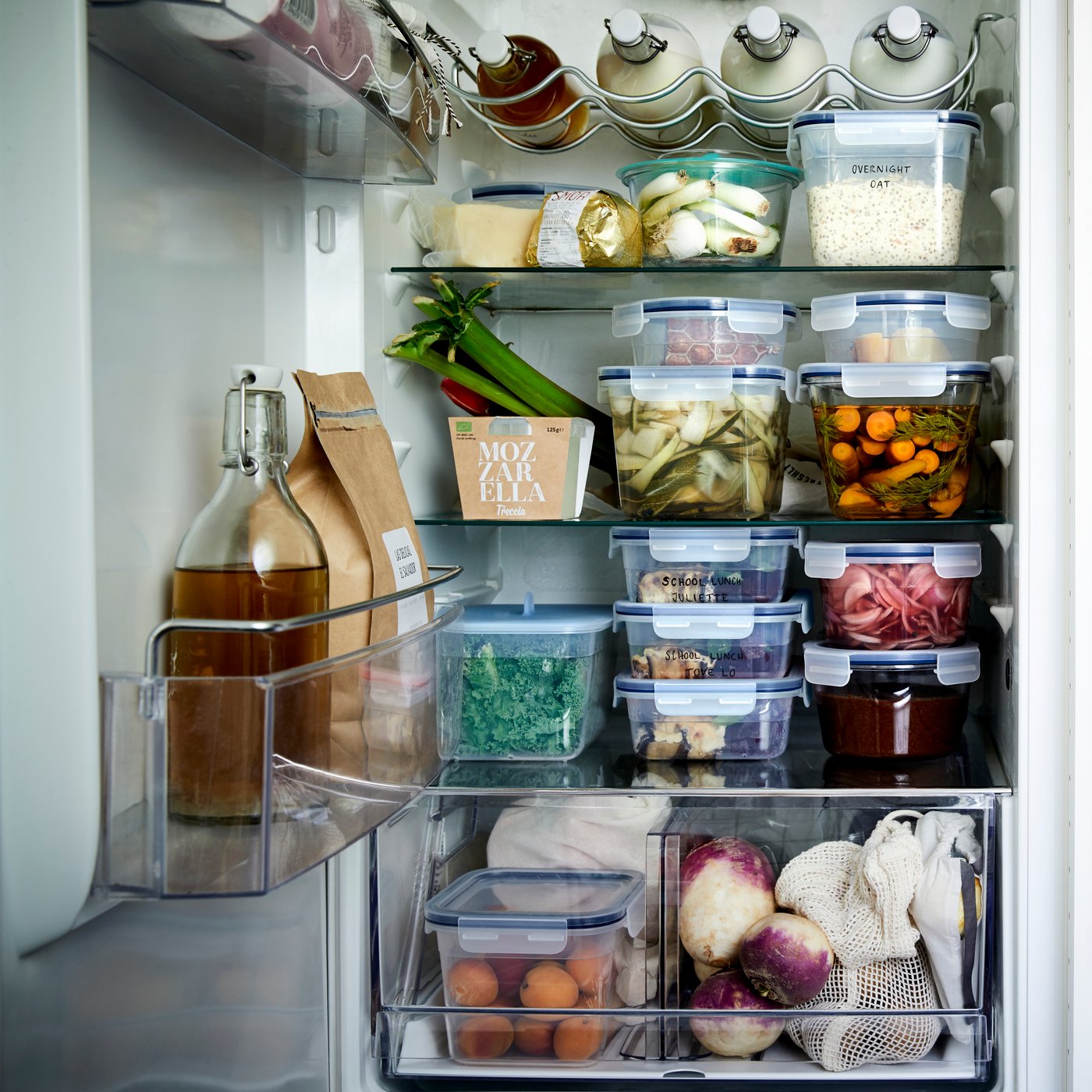 An open refrigerator is filled with glass bottles and IKEA 365+ plastic food storage containers full of food.