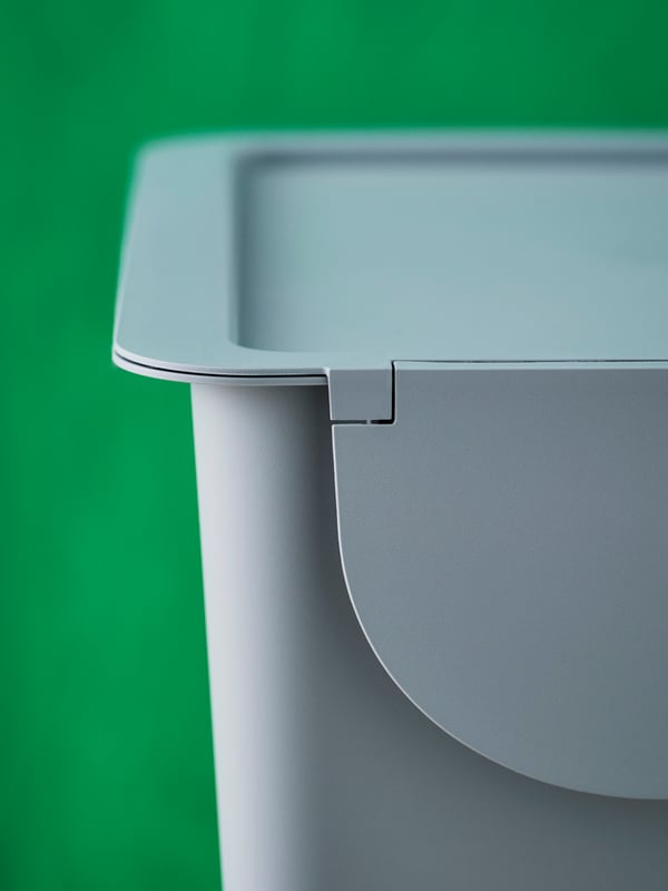 A close-up of a light grey-blue TRÖSKNING bin with lid against a green background.