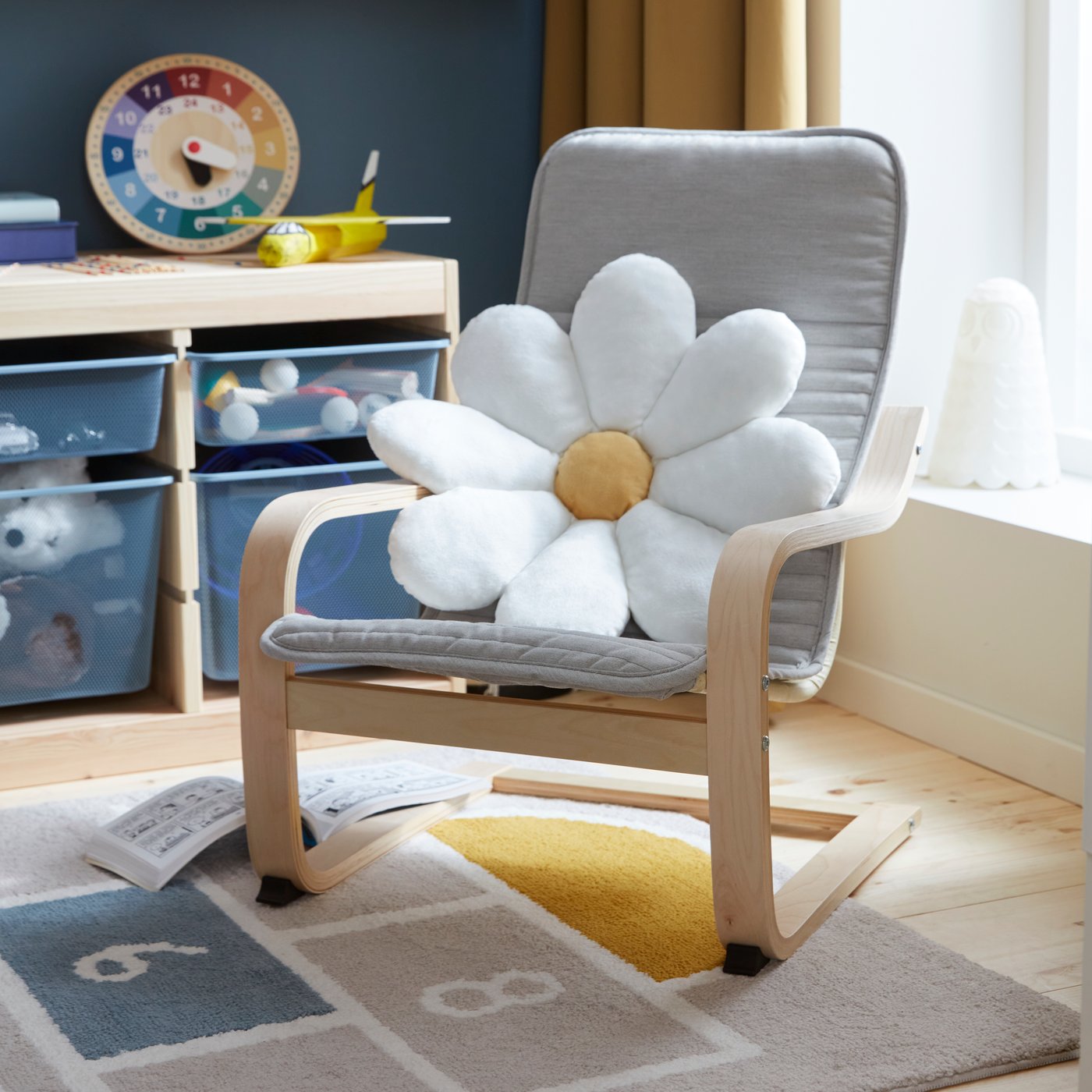 A daisy cushion is placed in a POÄNG children’s armchair in beige standing by a window in a children’s room with storage.