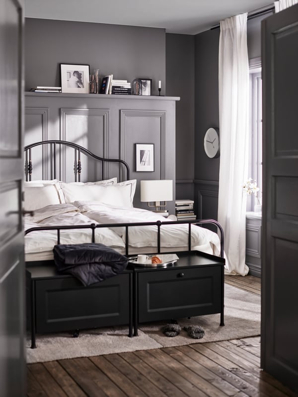 Twee zwarte KORNSJÖ opbergkisten en een bed met een metalen bed opgemaakt met wit/donkergrijs beddengoed op een lichtgrijs tapijt.
