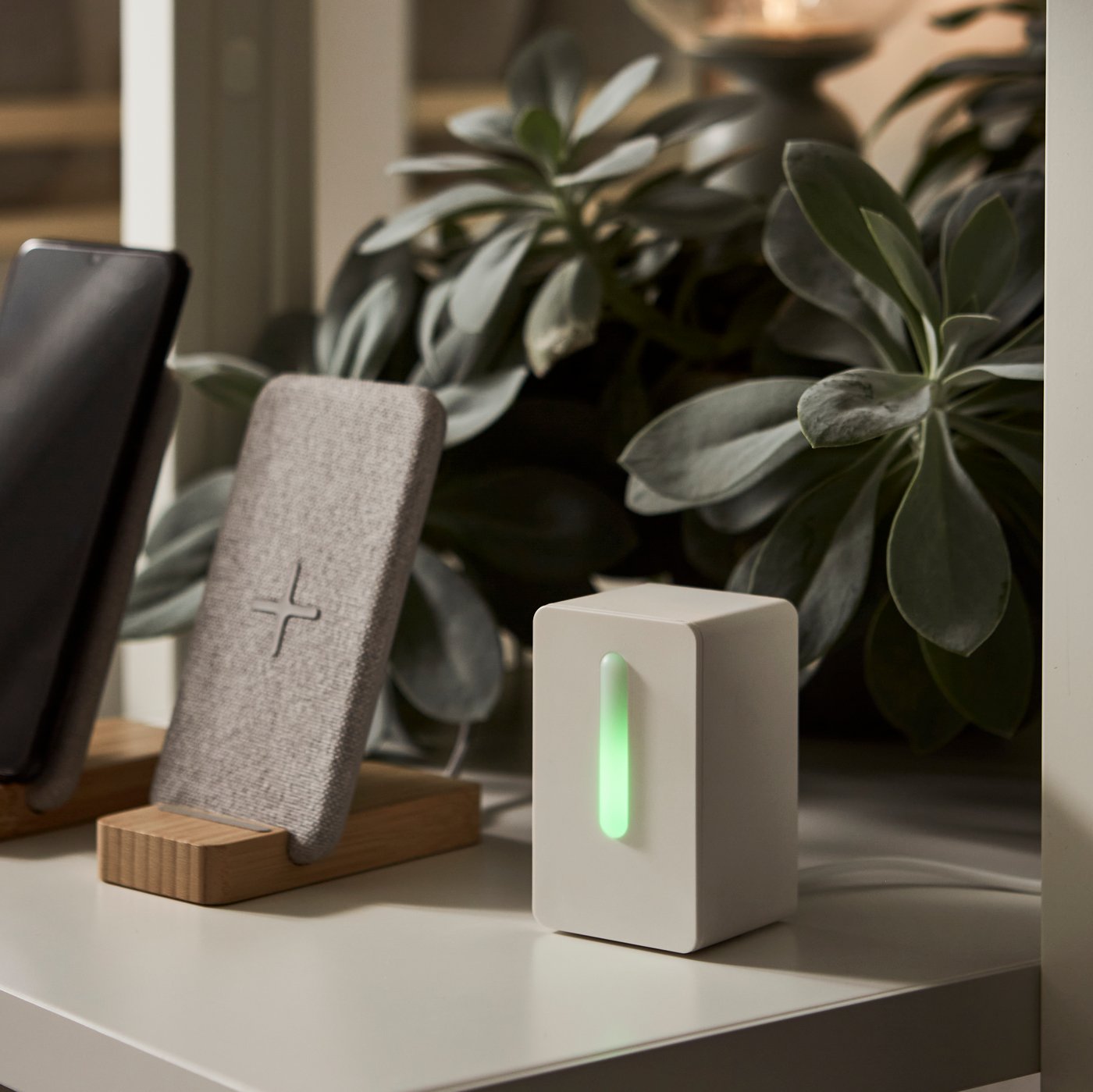 A VINDRIKTNING air quality sensor on a grey surface in front of a plant and beside a bamboo NORDMÄRKE wireless charger.