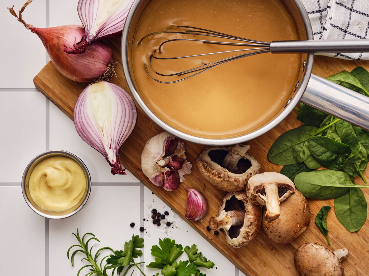På et skærebræt med hvidløg, svampe, krydderurter og skalotteløg er der et piskeris i en kasserolle med ALLEMANSRÄTTEN flødesaucemix.