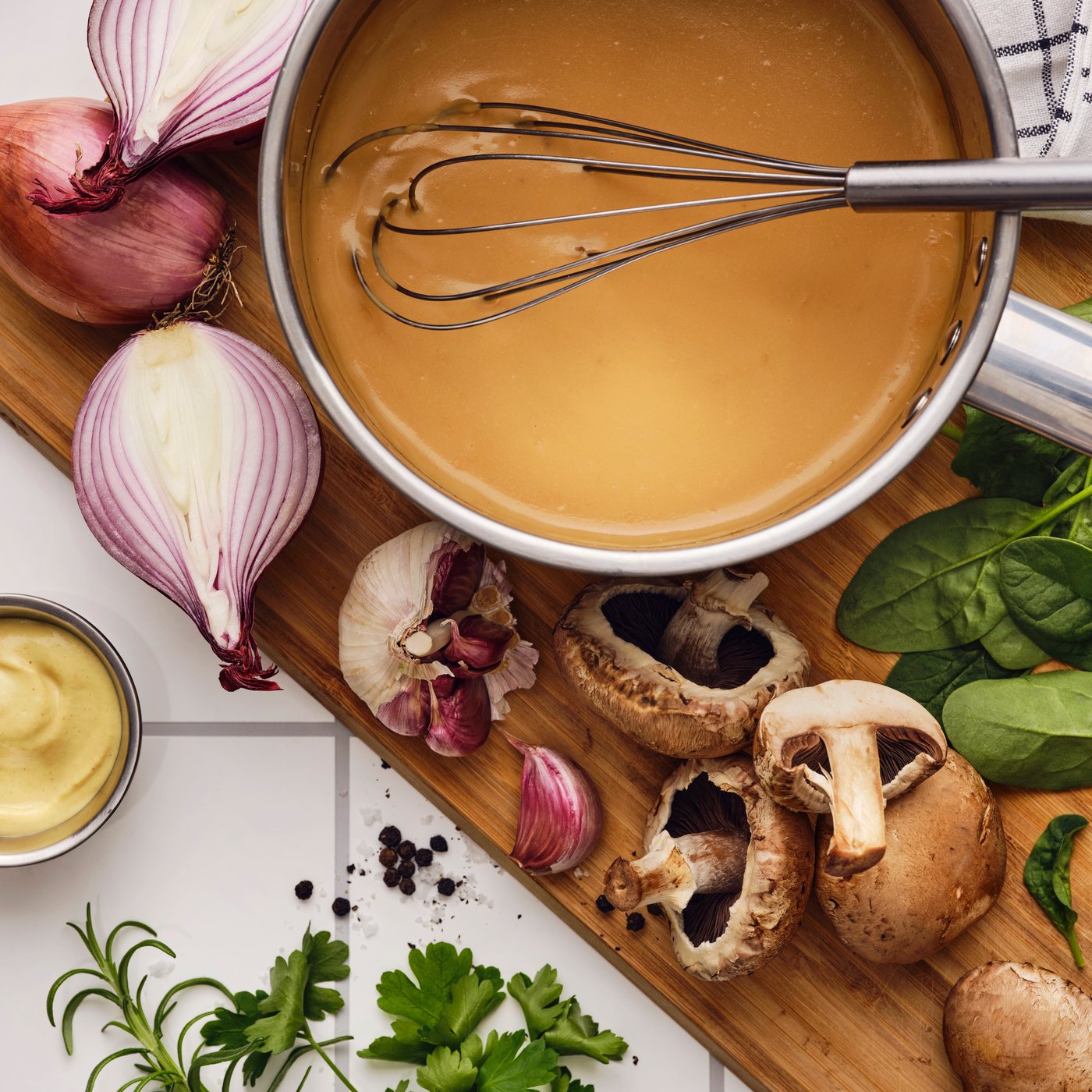 On a cutting board with garlic, mushrooms, herbs and shallots, a whisk is in a saucepan with ALLEMANSRÄTTEN savory sauce mix.
