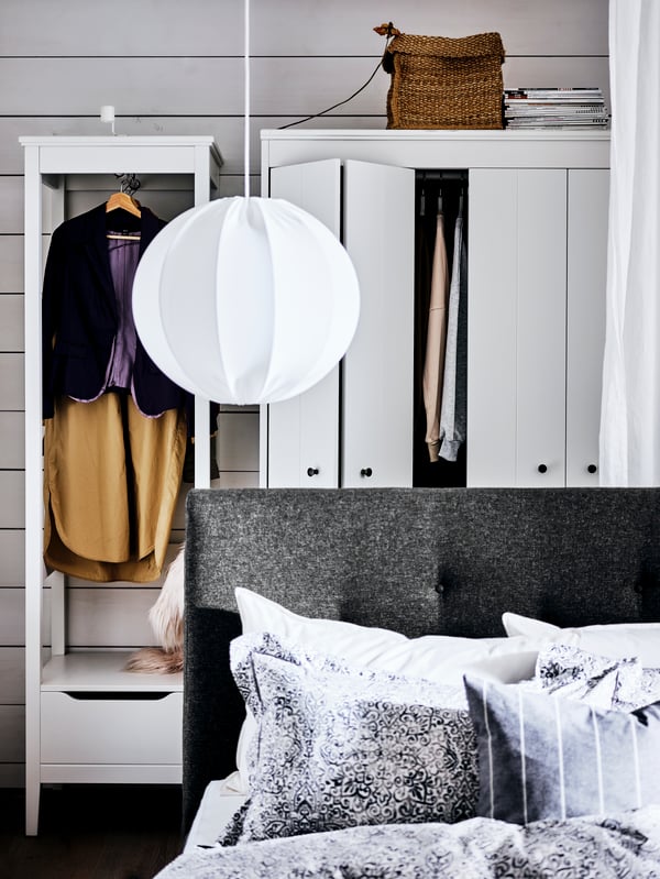 A grey IDANÄS upholstered bed with a pendant lamp above it, in front of two white wardrobes with hanging clothes on show.