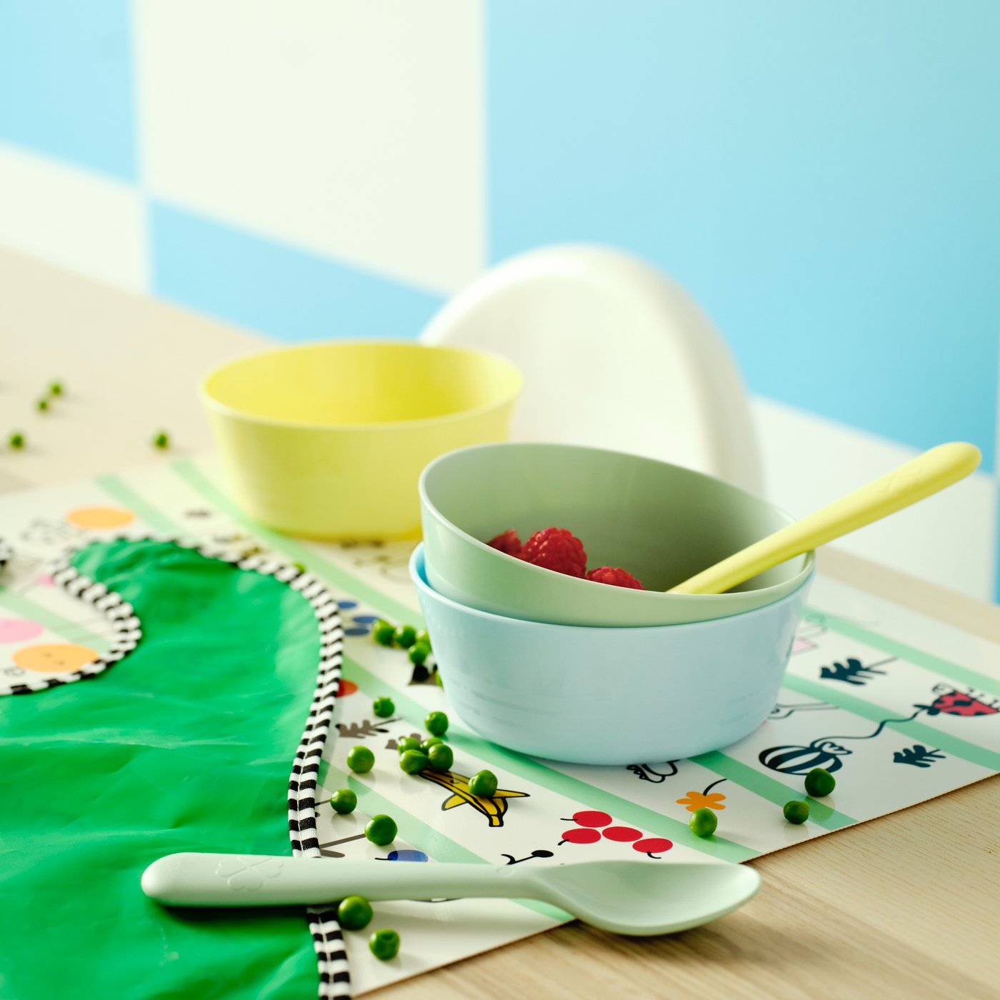 Yellow, green and blue KALAS tableware with raspberries and peas on a GRÖNFINK place mat, set against colourful decor.