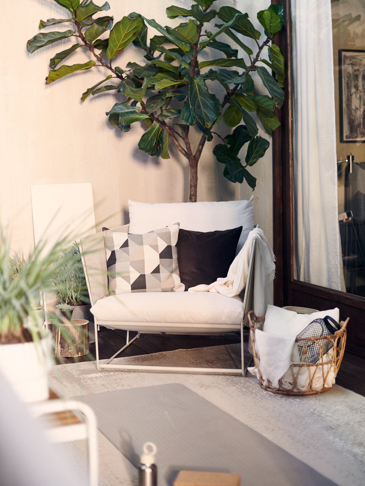 Un sillón HAVSTEN decorado con cojines, rodeado de plantas en una terraza soleada, con una canasta de mantas a un costado.