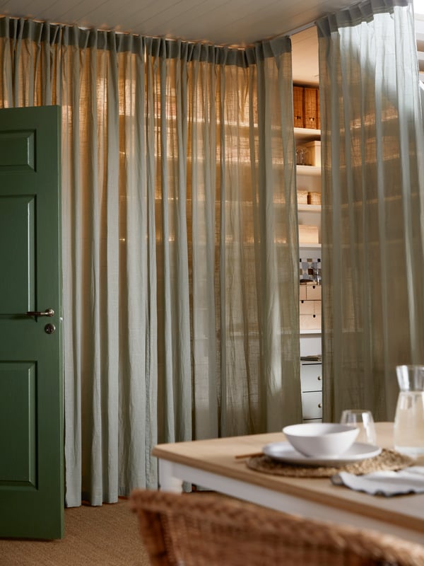 A corner of a room with VIDGA track set and SILVERLÖNN sheer curtains pulled closed hiding the wall shelves and storage.