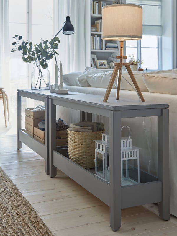 Two grey console tables are placed behind a sofa and serve as decorative room dividers. Two table lamps are placed on top.