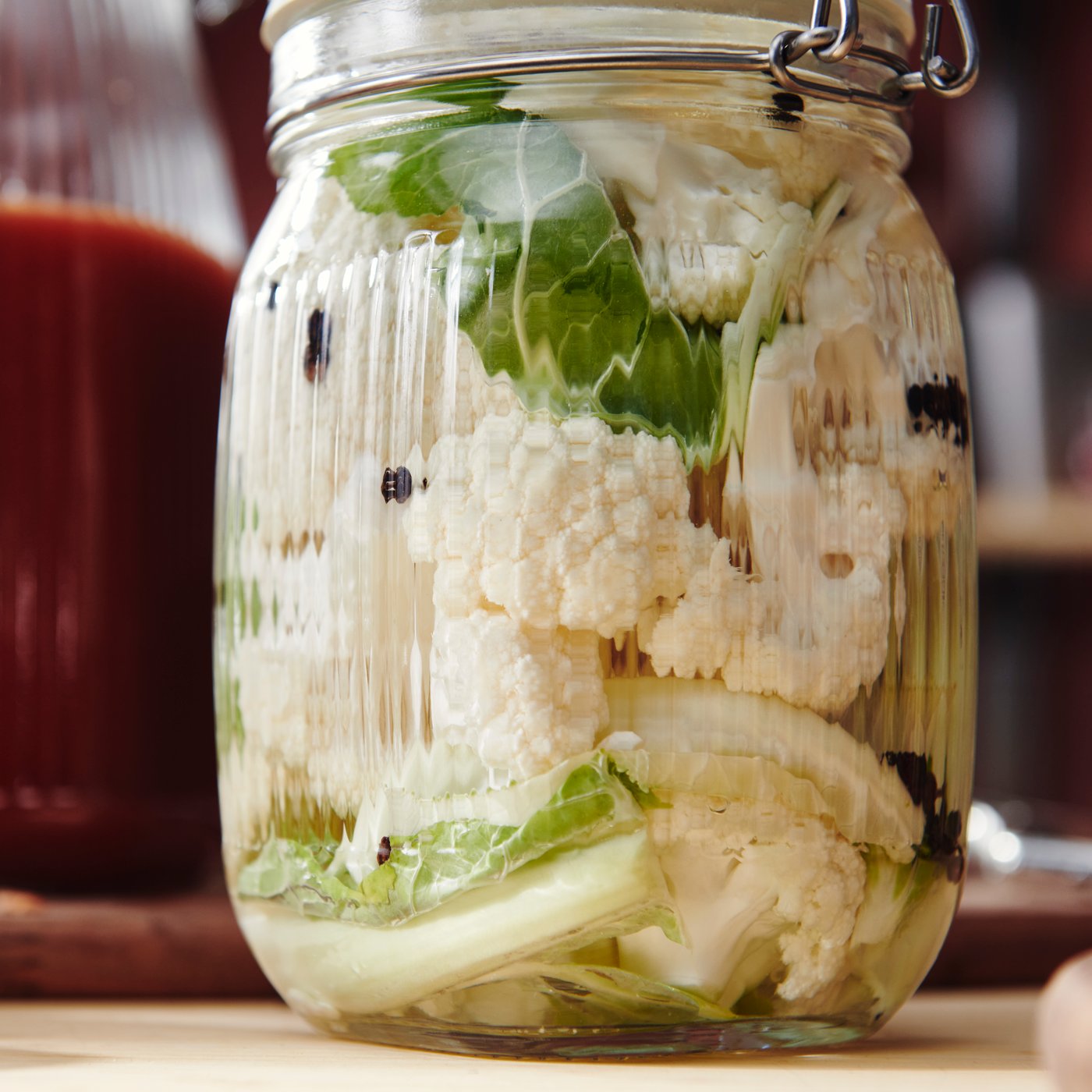 A clear glass KÖSSEBÄR jar with lid, filled with cauliflower and other vegetables, sits on a wooden worktop. 