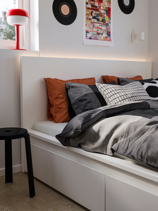 Close-up of a white MALM bed with drawers, BRUNKRISSLA bedding, a black stool by the side and a red lamp in the window.