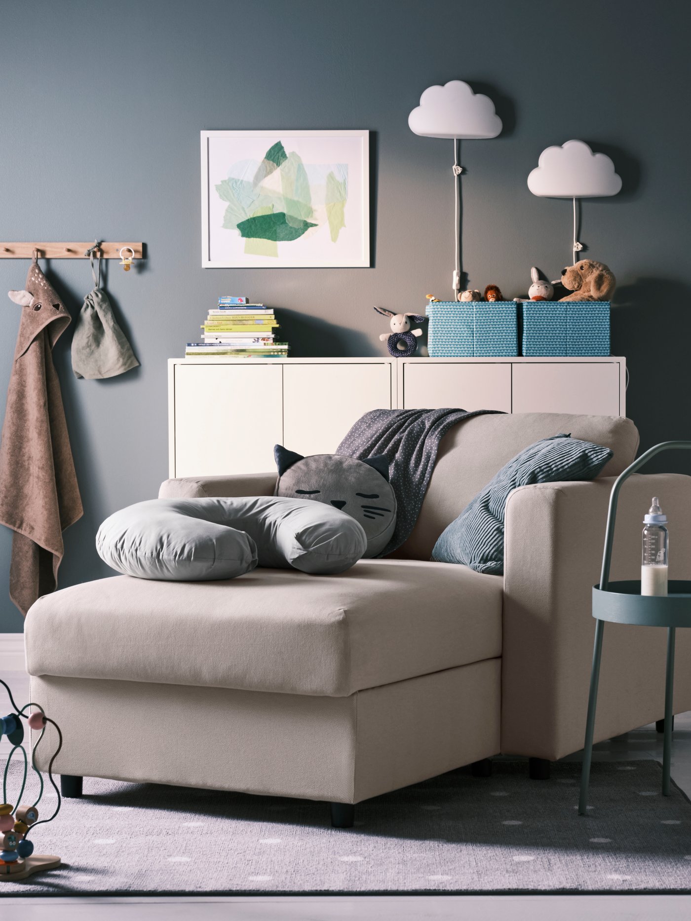 Una habitación con un chaise longue VIMLE beige y sobre él un cojín de lactancia LEN y otros cijines. Detrás un buffet EKET con lámparas de mesa y adornos.