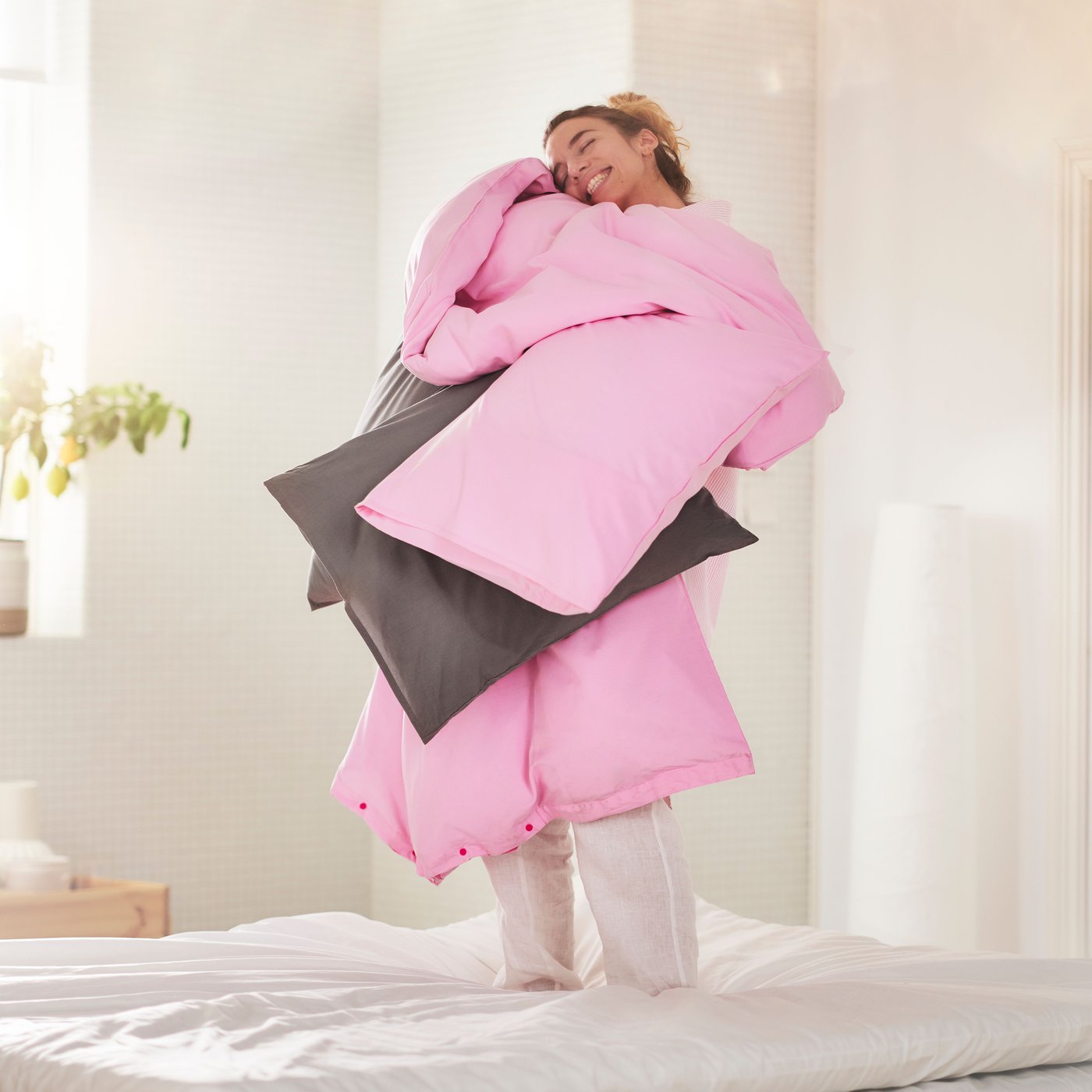 Personne souriante chargée d’une couette et d’oreillers habillés de linge de lit PILTANDVINGE rose pâle, dans une chambre lumineuse.