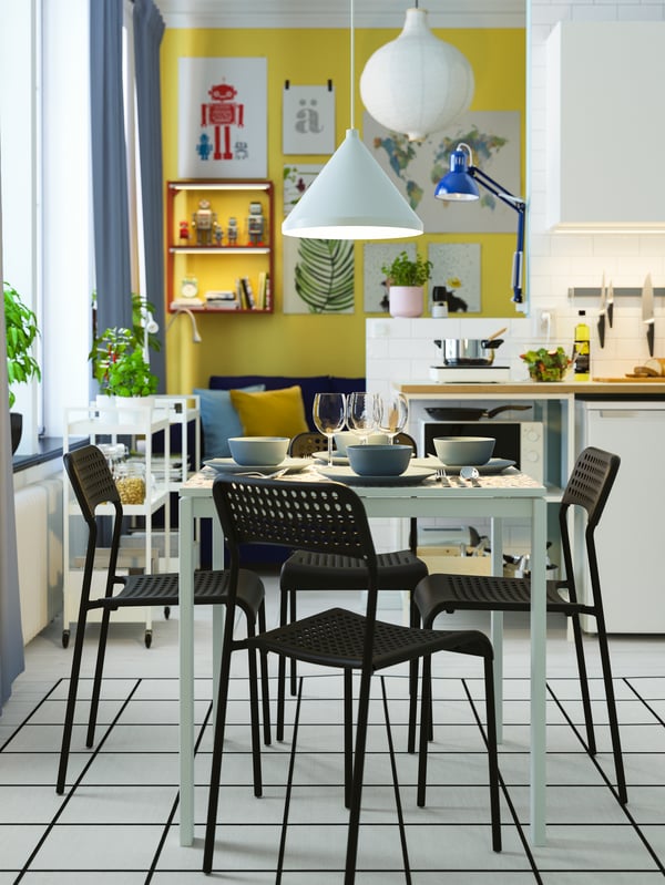 A white dining table with tableware, four black chairs, and a white pendant lamp turned on.