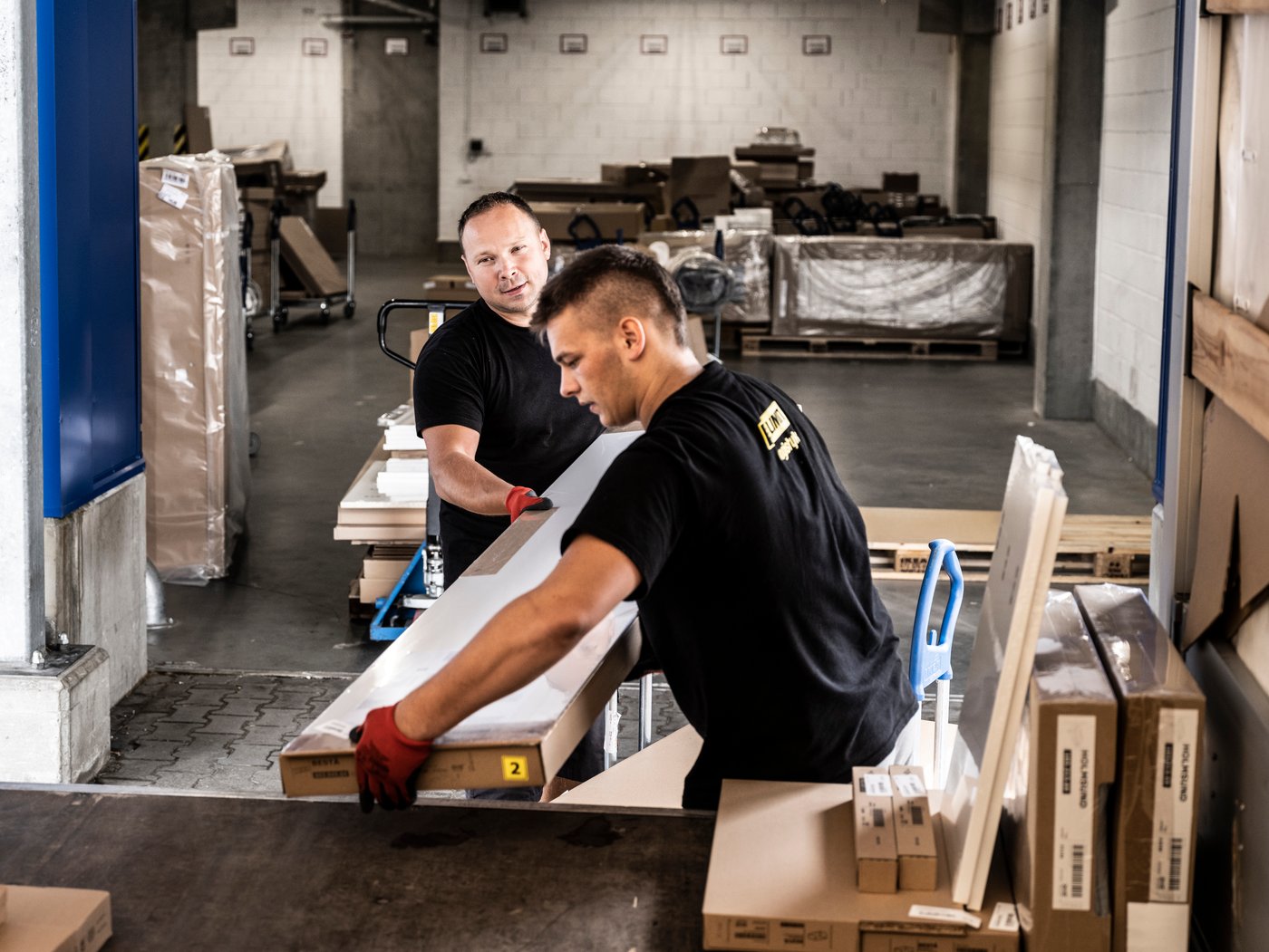 Dva muškarca u crnim majicama i crvenim radnim rukavicama utovaraju IKEA pljosnata pakovanja iz kolica u zadnji deo kamiona.