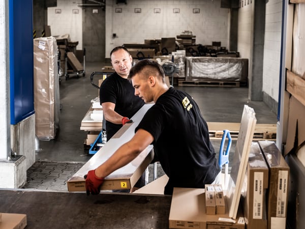 Two men wearing black t-shirts and red work gloves loading IKEA flatpacks from a trolley into the back of a truck.