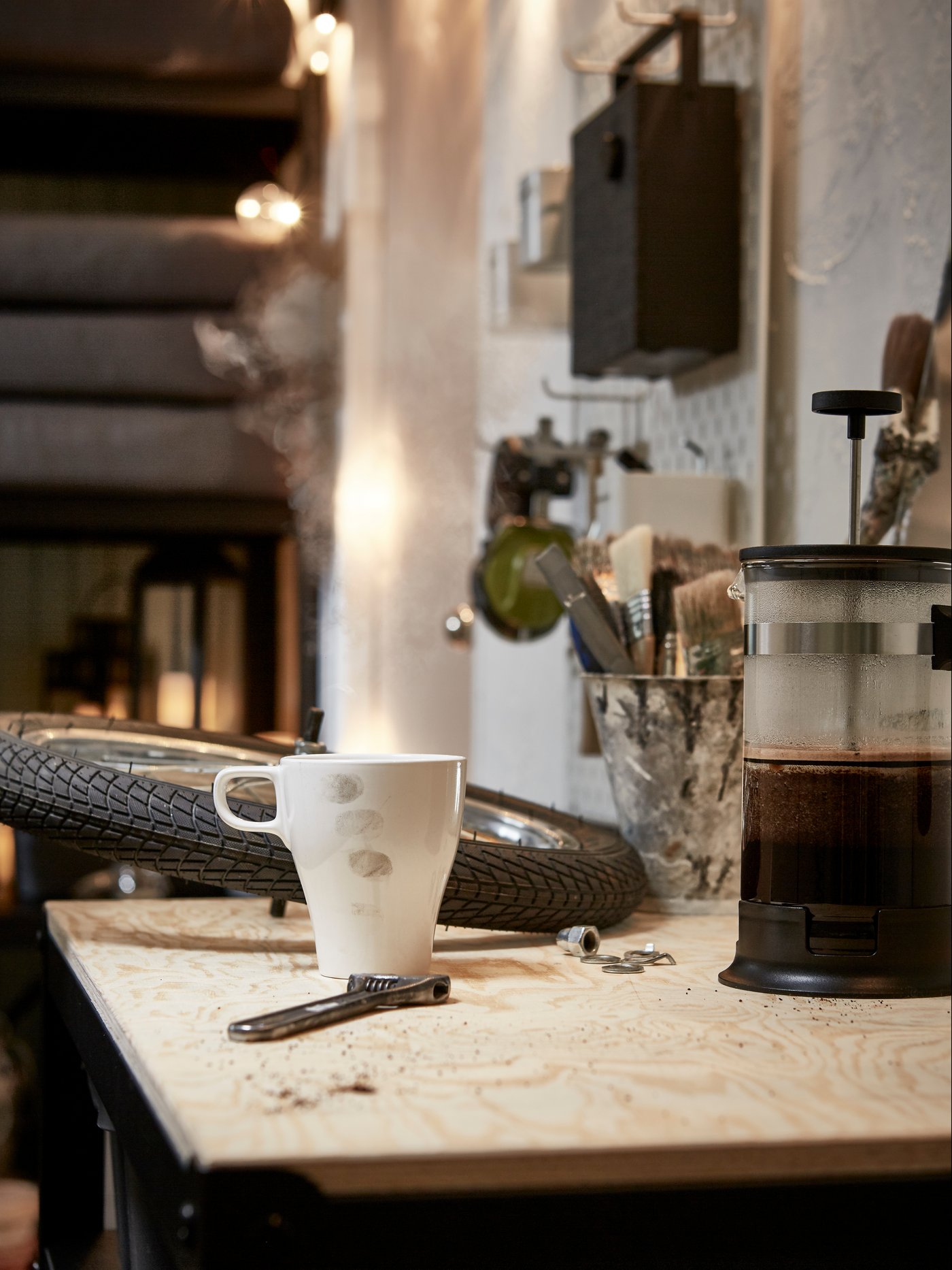 En benkeplate i et garasjerom med et sykkelhjul, en skiftenøkkel, et hvitt FÄRGRIK steintøyskrus og en dampende kaffetrakter i glass.
