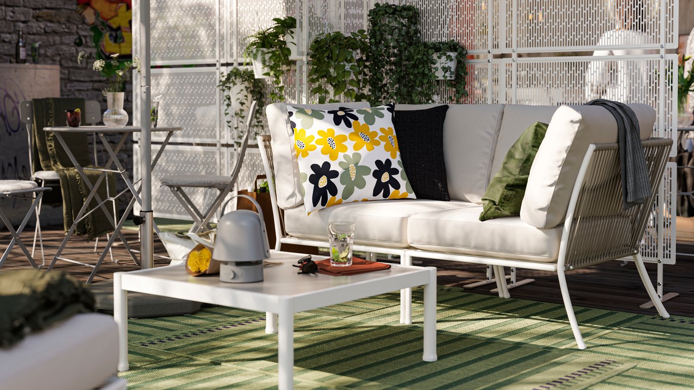 An outdoor lounge area with a two-seat SEGERÖN outdoor sofa and extra cushions. Near it are a cafe table and a coffee table.