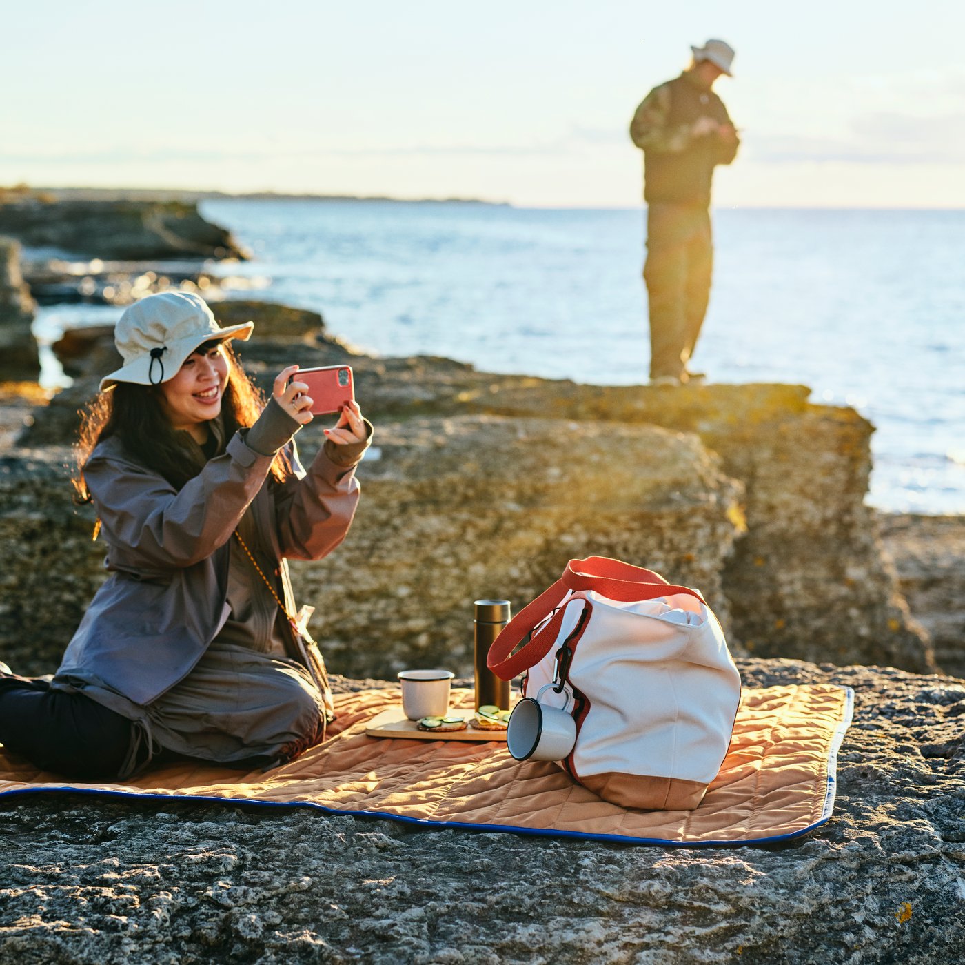Person sitting on a SOLUPPGÅNG picnic blanket near rocky shore, surrounded by outdoor gear including a large bag.