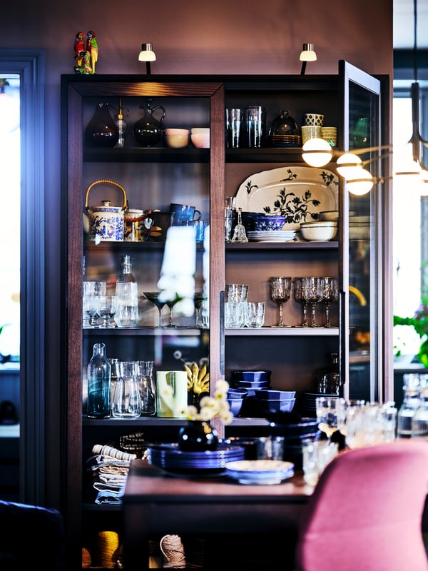 A brown REGISSÖR glass-door cabinet in a dining area with one door ajar housing a collection of crockery and glassware.