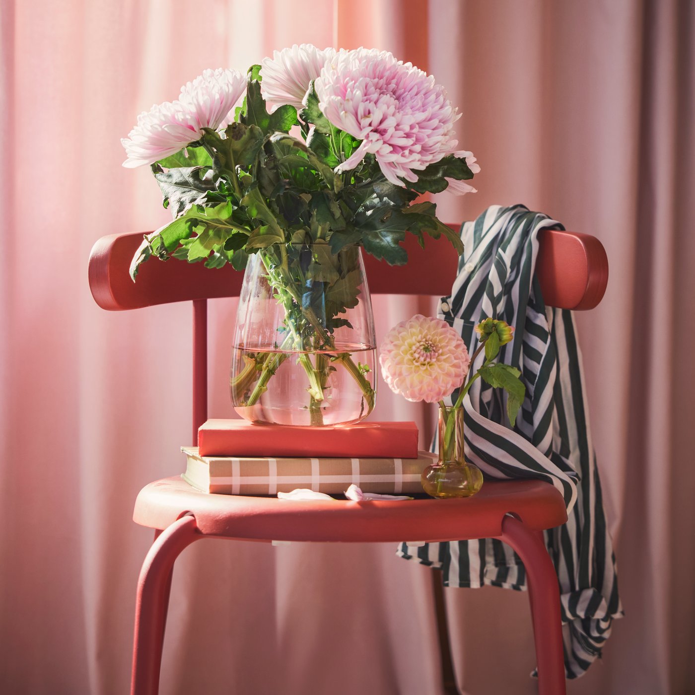 A living room features a light pink BERÄKNA vase made of glass filled with large pink flowers on a red chair beside books.
