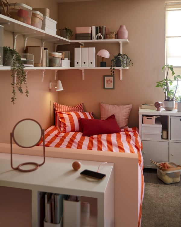 A small room with a white KALLAX desk by the foot end of a bed with bed linen in bright colours and wall shelves above it.