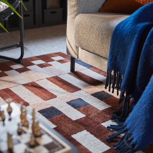 Una sala con una alfombra MARKBLÅMME de pelo corto y diseño de cuadros multicolor, colocada debajo de un sofá y junto a un juego de ajedrez.