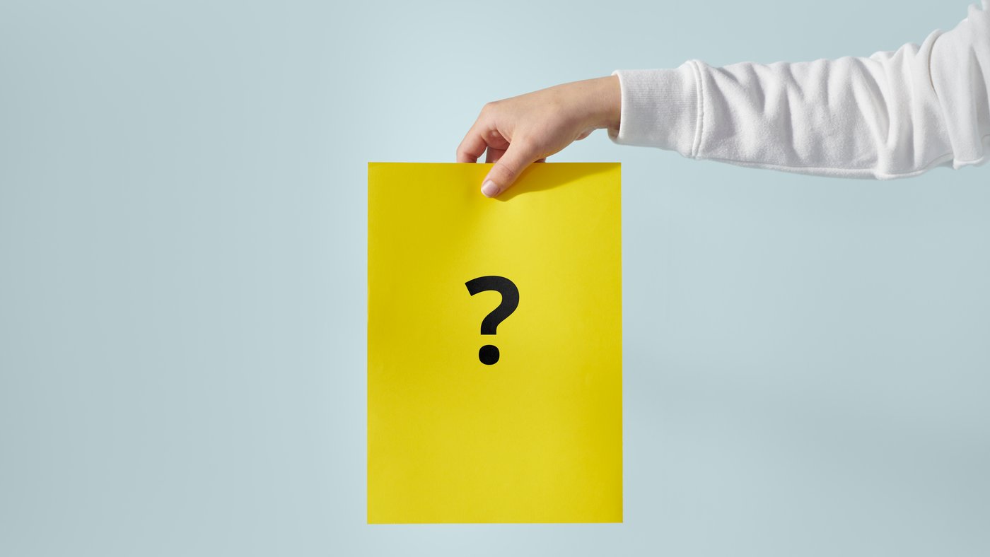 Une main tient une feuille A4 jaune avec un point d'interrogation noir.