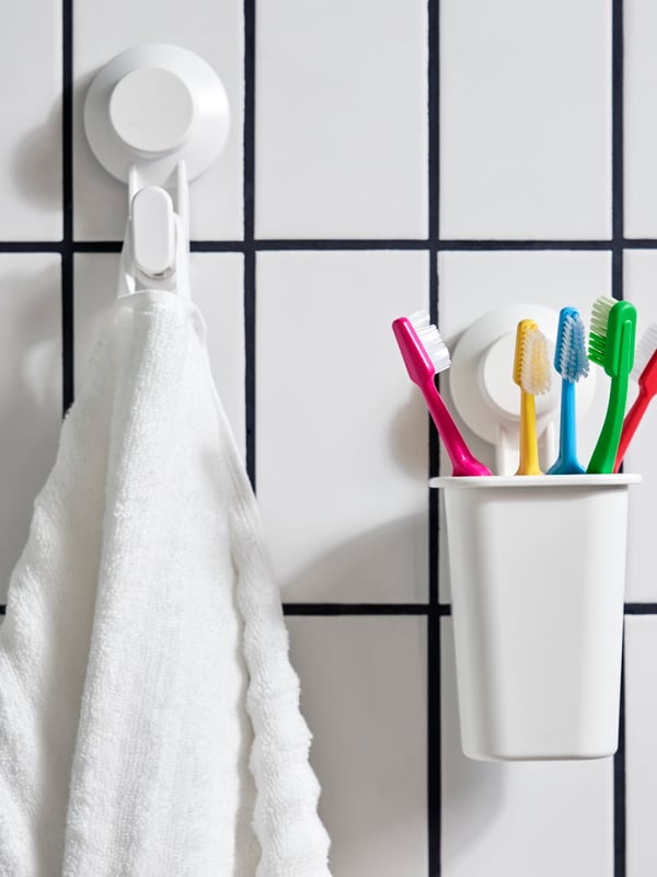 TISKEN wall suction cup holder mounted on a wall with a few multicoloured toothbrushes inside next to a TISKEN wall suction hook with a towel hanging on it.