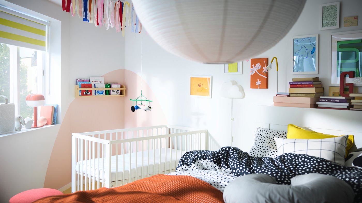 Chambre à coucher avec grand lit à l’avant-plan et lit bébé à l’arrière-plan, plusieurs étagères avec des livres et de nombreux tableaux.