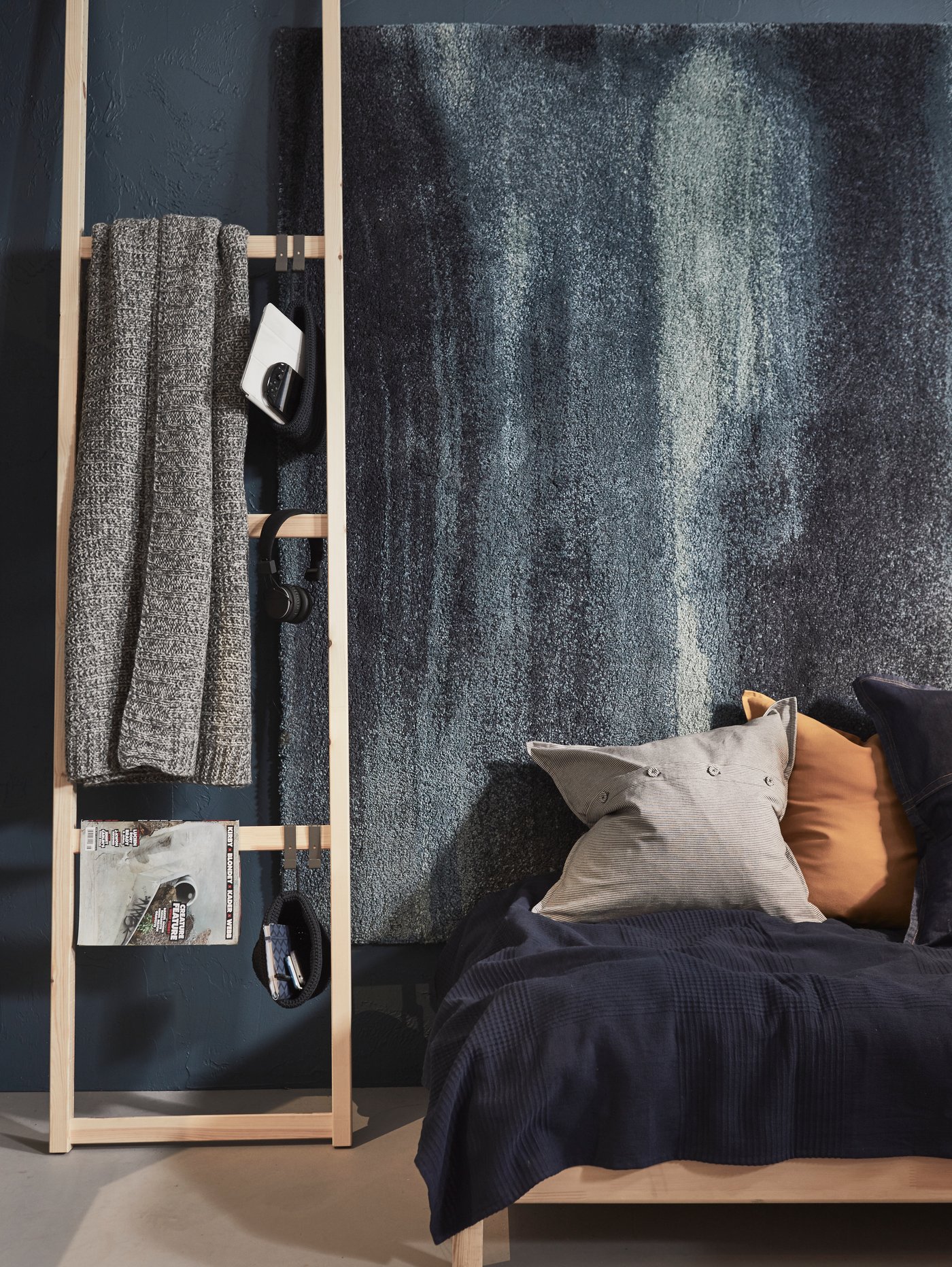 Un montant IVAR contre un mur de chambre à coucher avec un jeté gris, un magazine, des paniers NORDRANA et des écouteurs suspendus.