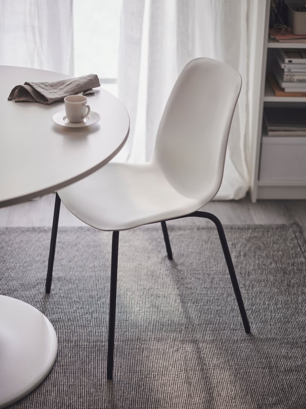 A light room with a chair formed by a black SEFAST underframe and white LIDÅS seat shell, by a round table, on a TIPHEDE rug.