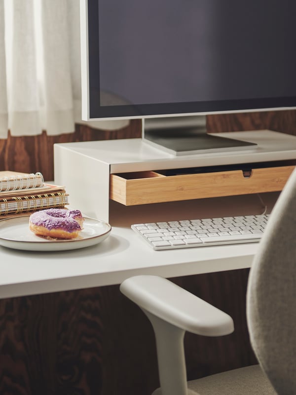 A workspace with a doughnut on a side plate placed by a white ELLOVEN monitor stand on a white TROTTEN sit-stand desk.
